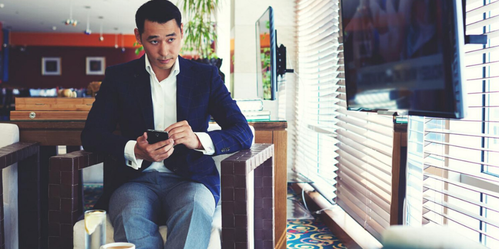A man in a navy blazer sits in a modern lounge, holding a smartphone and looking to the side, as if preparing for customer service conversations. Sunlight streams through window blinds, with a large TV nearby and a drink on the table before him.