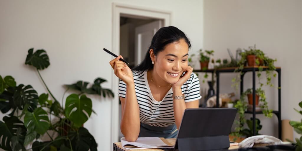 A woman smiles while looking at a tablet on a desk, holding a pen. She is surrounded by notebooks and plants in a bright, cozy room, possibly preparing to create a comprehensive report or review customer satisfaction survey results.