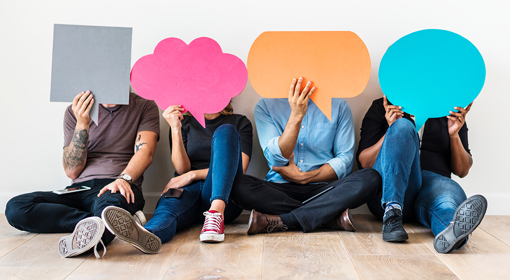 Four people sit on the floor against a wall, each holding a large, colorful speech or thought bubble cutout in front of their faces, symbolizing conversational feedback and real-time customer feedback that can drive business growth.