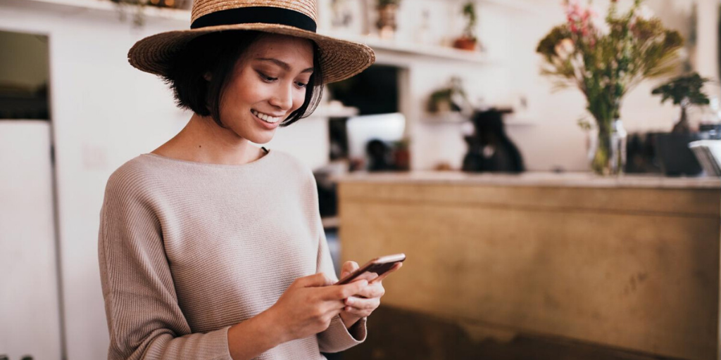 A woman wearing a straw hat and beige sweater smiles while looking at her smartphone in a cozy, well-lit café—perhaps checking tips from CX Leaders to boost her CSAT, with flowers and shelves in the background.