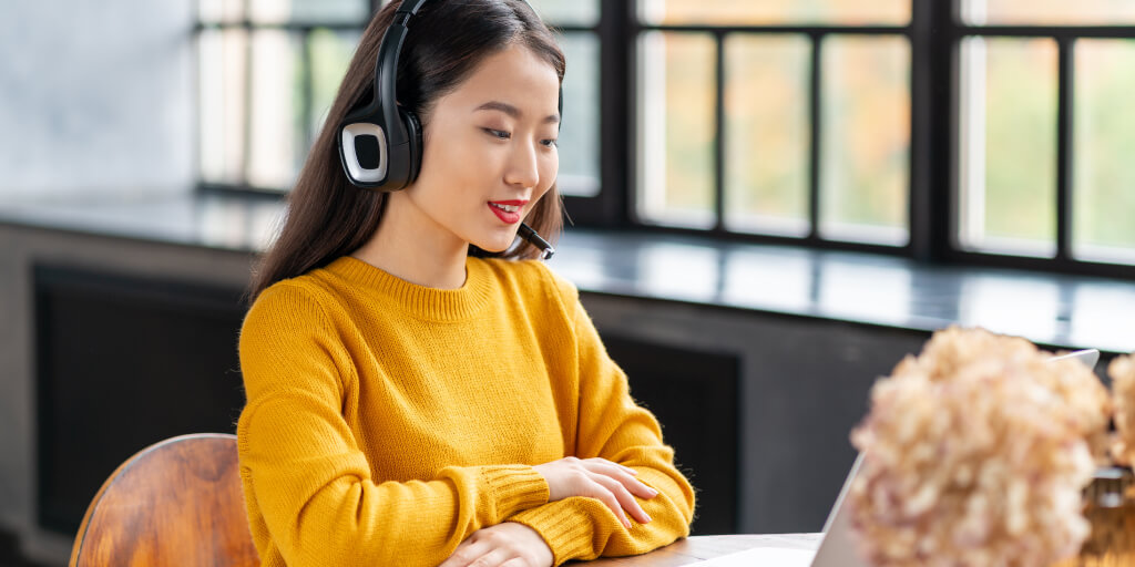 A woman wearing a yellow sweater and a headset sits at a desk, looking at a laptop screen and speaking about boosting agent productivity, with large windows and blurred flowers in the background.