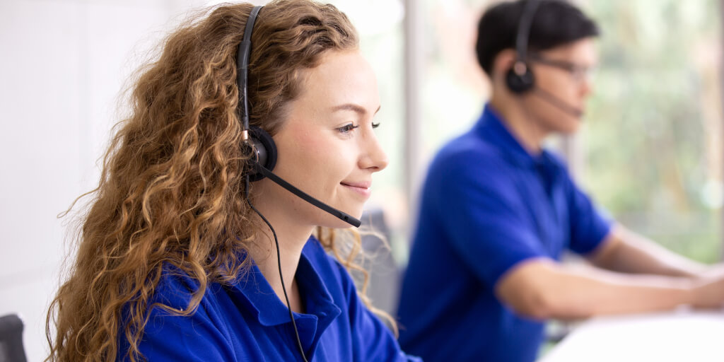 A woman with curly hair wearing a headset and blue shirt sits at a desk, smiling while talking. Beside her, a man in a blue shirt and headset works at a computer as they strive to meet call center benchmarks for service quality.