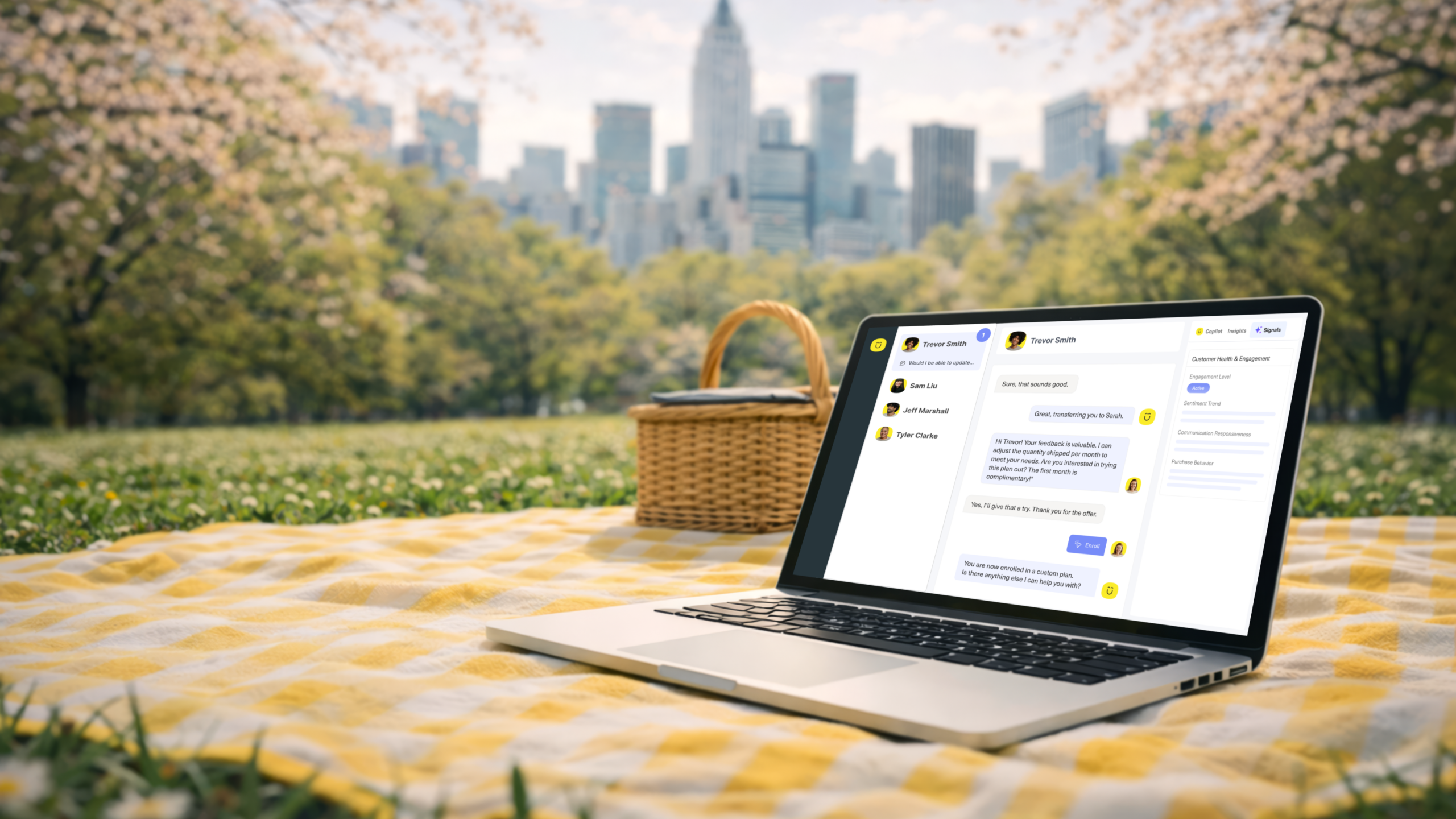 A laptop sits on a yellow picnic blanket in a park, displaying the Kustomer chat interface. Nearby, a wicker basket rests under a bright sky with trees and a city skyline&mdash;hinting at exciting Summer 2025 product release updates.