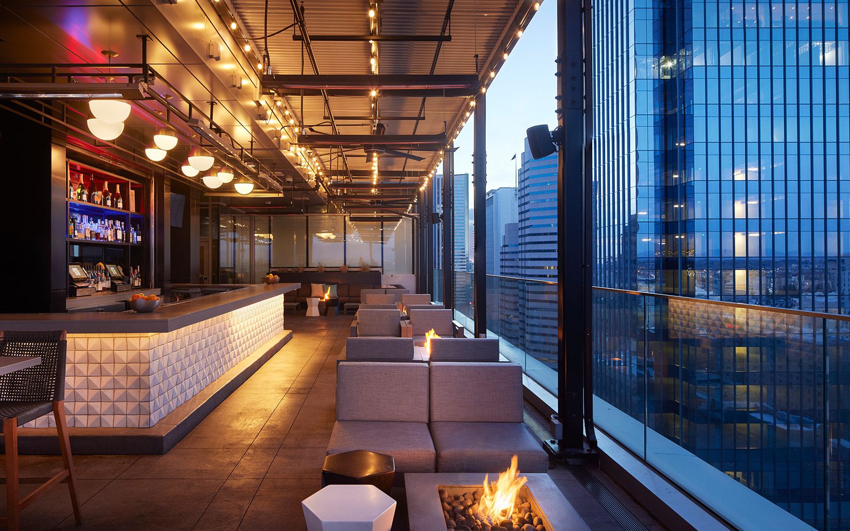 Modern rooftop bar at dusk, with cozy seating, fire pits, and string lights. City skyscrapers shine through glass railings while the stylish, illuminated bar creates a warm vibe&mdash;perfect for a relaxed Kustomer AI demo in an inviting setting.