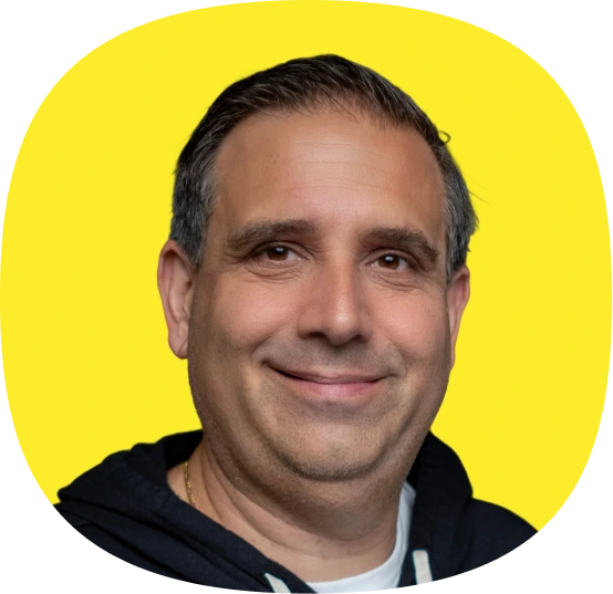 A middle-aged man with short dark hair smiles at the camera, exuding confidence. He wears a dark hoodie over a white shirt, set against a solid bright yellow backdrop—perfect for highlighting Kustomer Winter Quarterly AI Innovations.