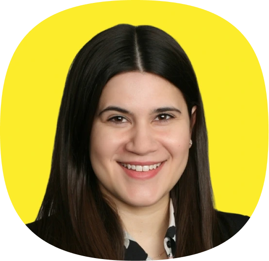 A woman with straight, dark brown hair and fair skin smiles at the camera. She is wearing a black jacket and a patterned shirt, standing against a bright yellow background that radiates energy—perfect for Kustomer Winter Quarterly AI Innovations.