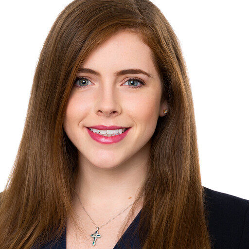 A young woman with long, straight brown hair and blue eyes smiles at the camera. She is wearing a dark top and a silver necklace with a cross pendant. The background is white.