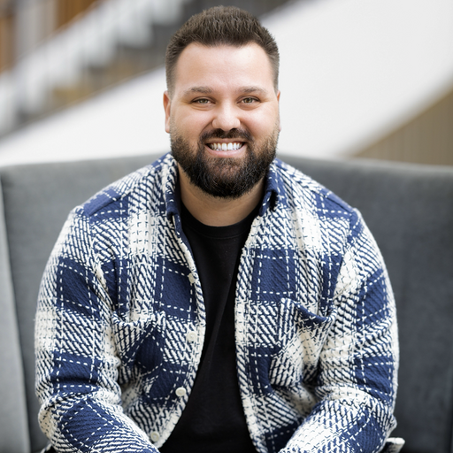 A man with a beard and short hair, wearing a blue and white plaid jacket over a black shirt, smiles while sitting on a gray chair in a modern indoor setting.