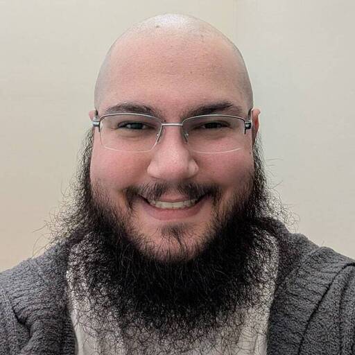 A smiling person with a bald head, glasses, and a full dark beard, wearing a gray sweater and a light-colored shirt, poses in front of a plain, light-colored background.