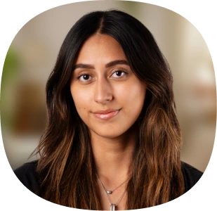 A young woman with long, wavy dark hair and brown highlights is looking at the camera with a slight smile—capturing the confidence inspired by Aplazo’s AI-Powered Customer Support with Kustomer AI. She wears a necklace and a dark top, indoors softly blurred behind her.