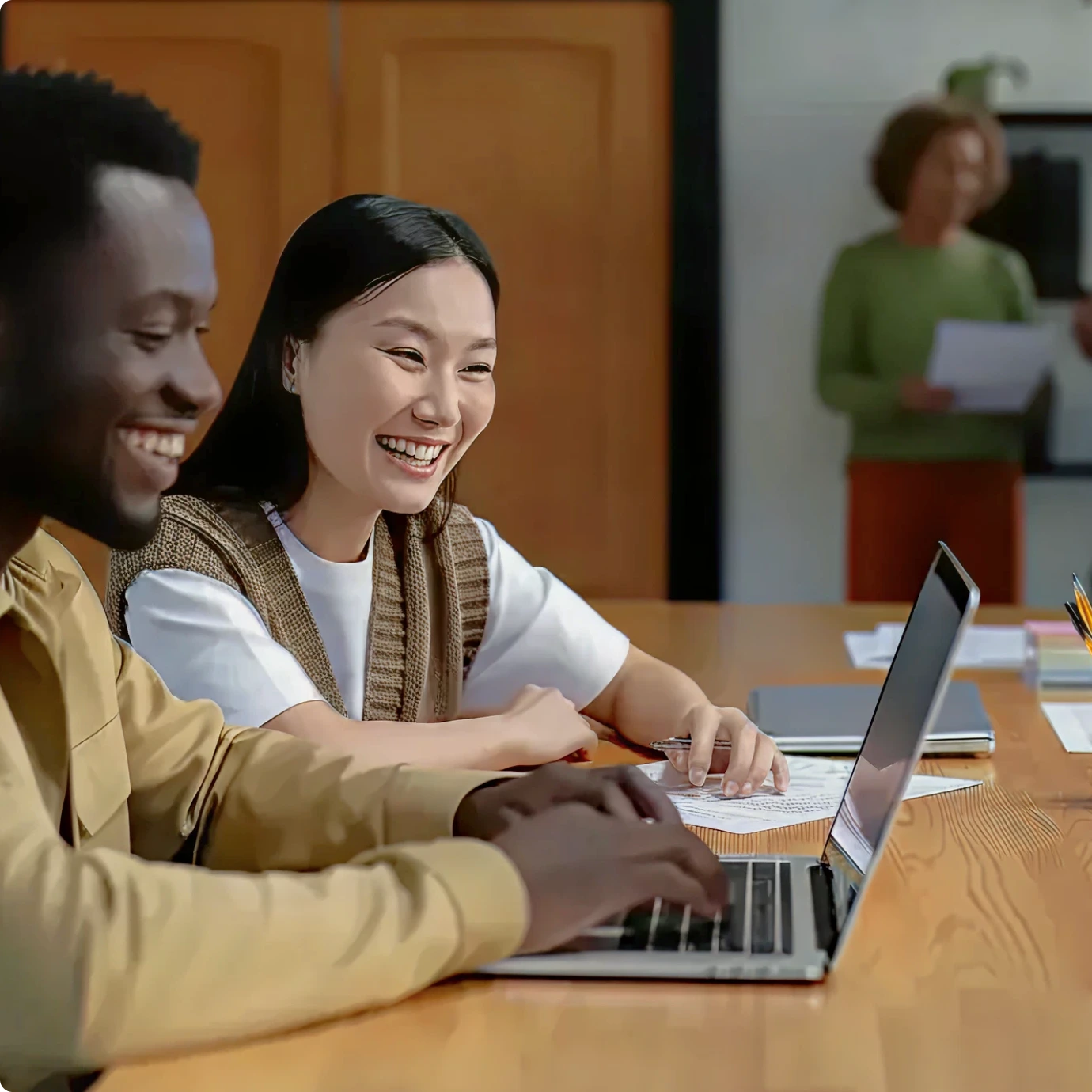 Two young adults, a man and a woman, smile and work together at a laptop in a classroom setting. A person stands in the background holding papers near a whiteboard.