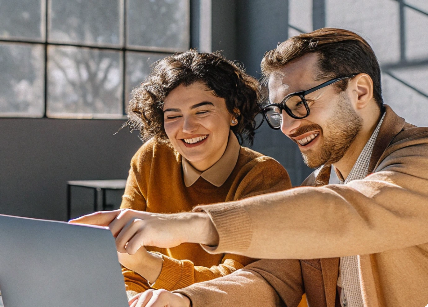 Two people smiling and sitting at a table, looking at a laptop screen together. Sunlight streams through large windows behind them, creating a bright and cheerful atmosphere.
