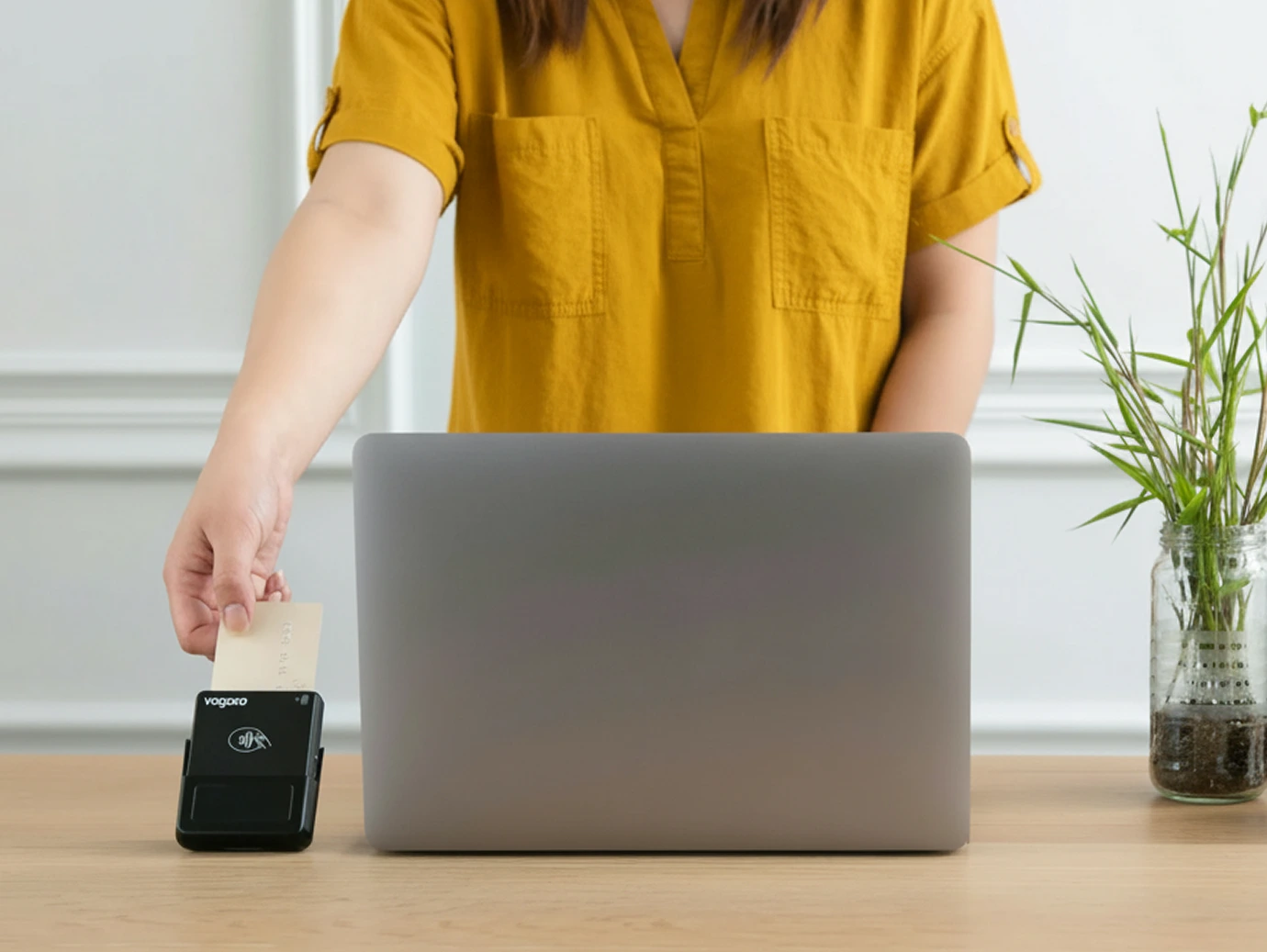 A person in a mustard yellow shirt swipes a card through a payment terminal next to a laptop on a wooden desk; a glass jar with green plants is also on the desk.