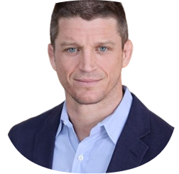A man with short brown hair and blue eyes wearing a light blue collared shirt and a dark navy blazer, posing in front of a plain white background.
